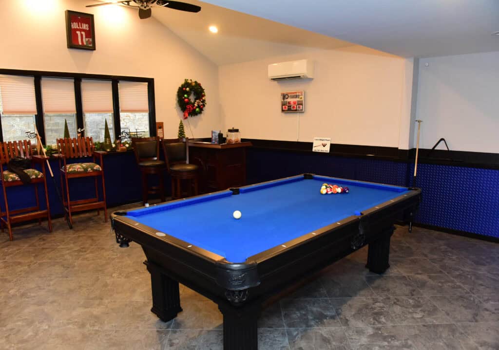 A game room with a blue pool table, cue stick on the wall, high chairs at a counter, holiday wreath on the window, framed sports jersey, and a small bar area in the corner.