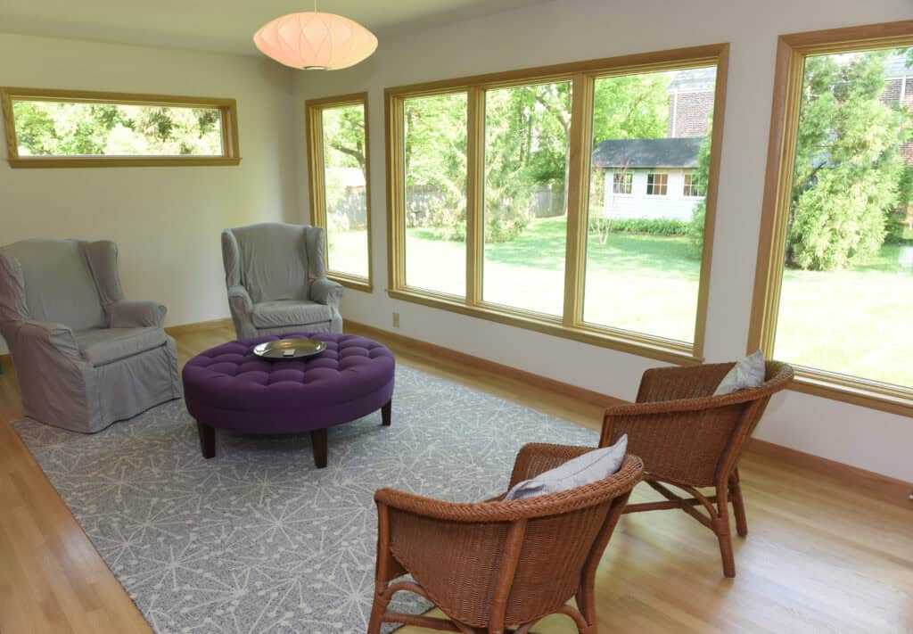 Bright living room with large windows, two gray armchairs, two wicker chairs, a round purple ottoman on a patterned rug, wood floors, and views of a green yard with trees and a white shed outside.