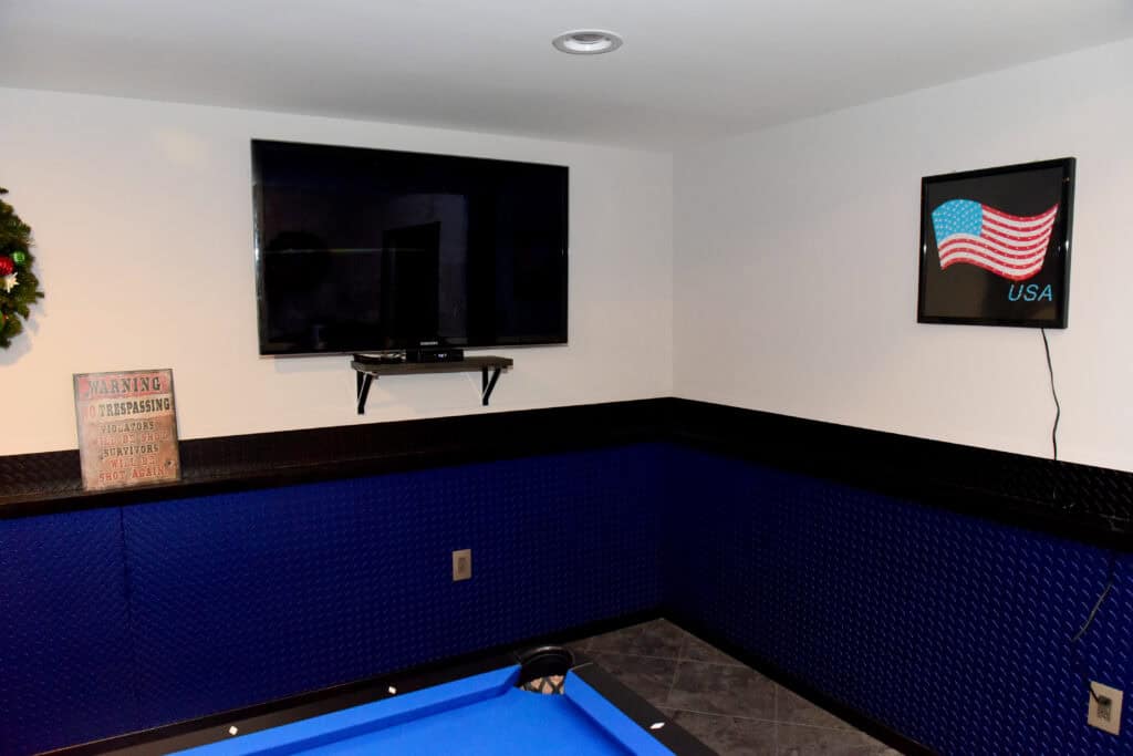 A room corner with a blue pool table, a wall-mounted TV above a shelf, a sign with a warning, and a glowing USA flag sign on the wall. The walls are white with blue paneling along the bottom.