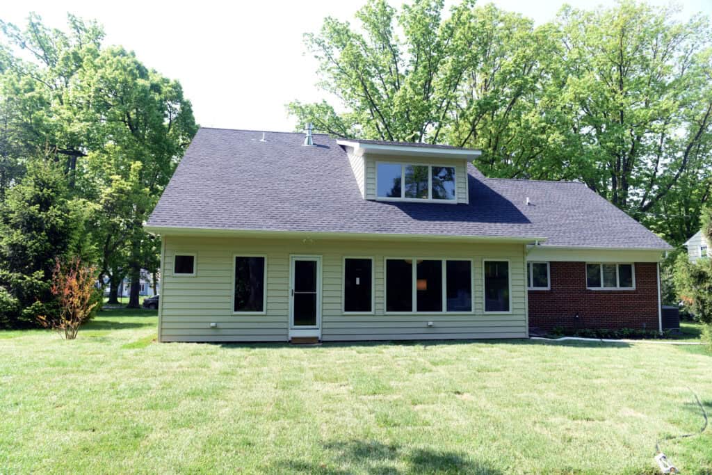 A house with light yellow siding and red brick, large windows, and an upper dormer, set in a grassy yard with green trees in the background on a sunny day.
