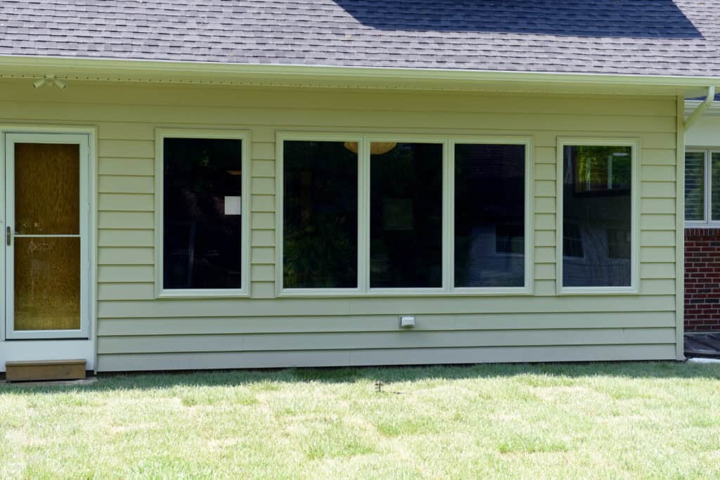 A beige house exterior with three large windows and a wooden door, all facing a green lawn. The siding is horizontal, and the roof has dark shingles. Reflections are visible in the window glass.