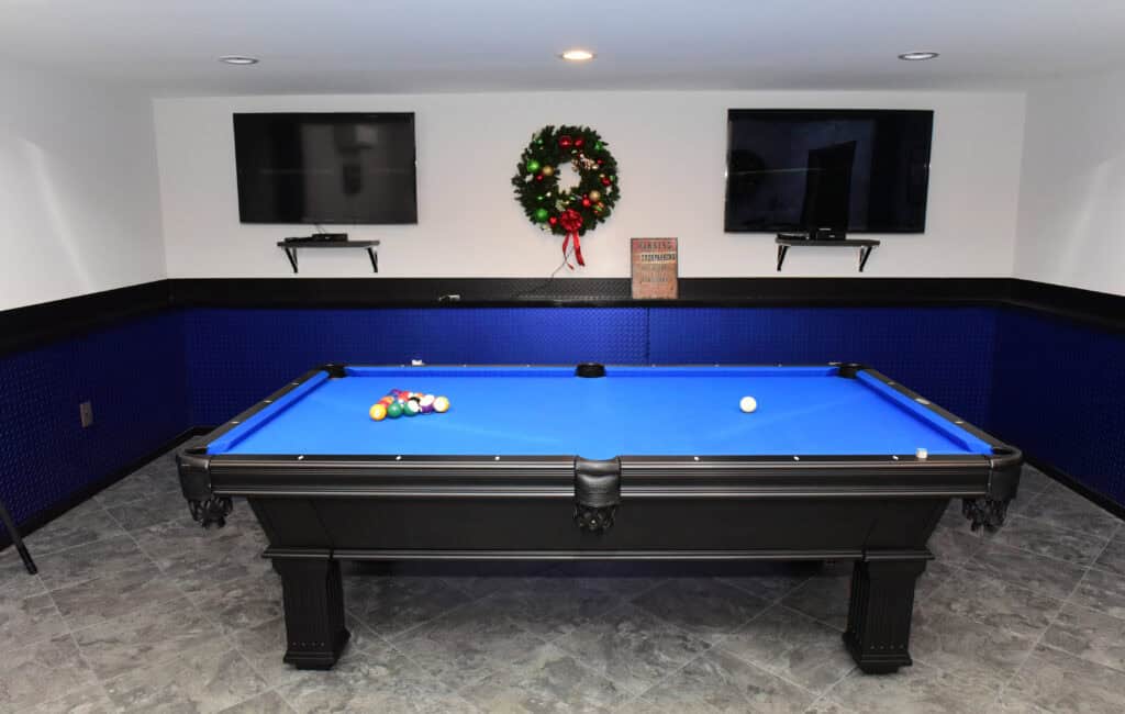 A blue pool table with racked balls is centered in a room with gray tile floors. Two wall-mounted TVs, a festive wreath, and a small sign are on the white wall behind the table.
