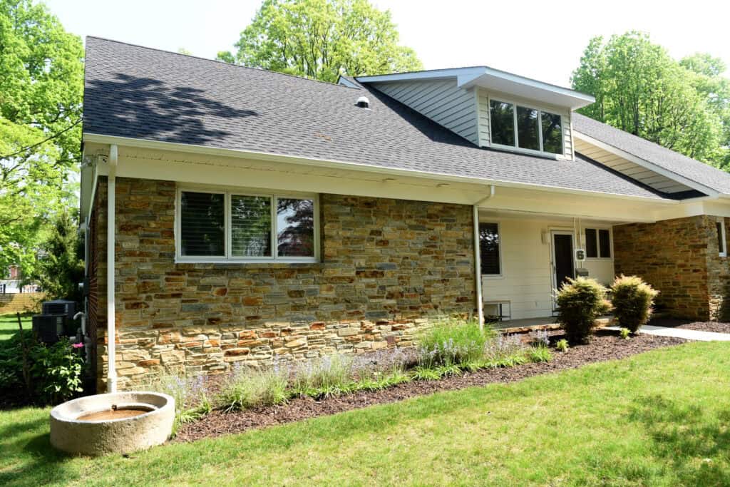 A modern house with stone and white siding, black shingle roof, and dormer window. There is a neatly maintained garden bed and a grassy yard with trees in the background.