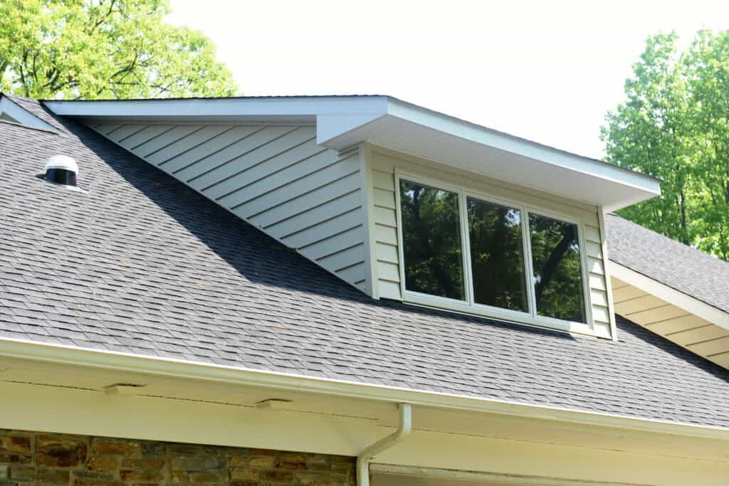 A close-up view of a house roof with gray shingles, a dormer window with white trim and horizontal siding, and green trees in the background on a sunny day.