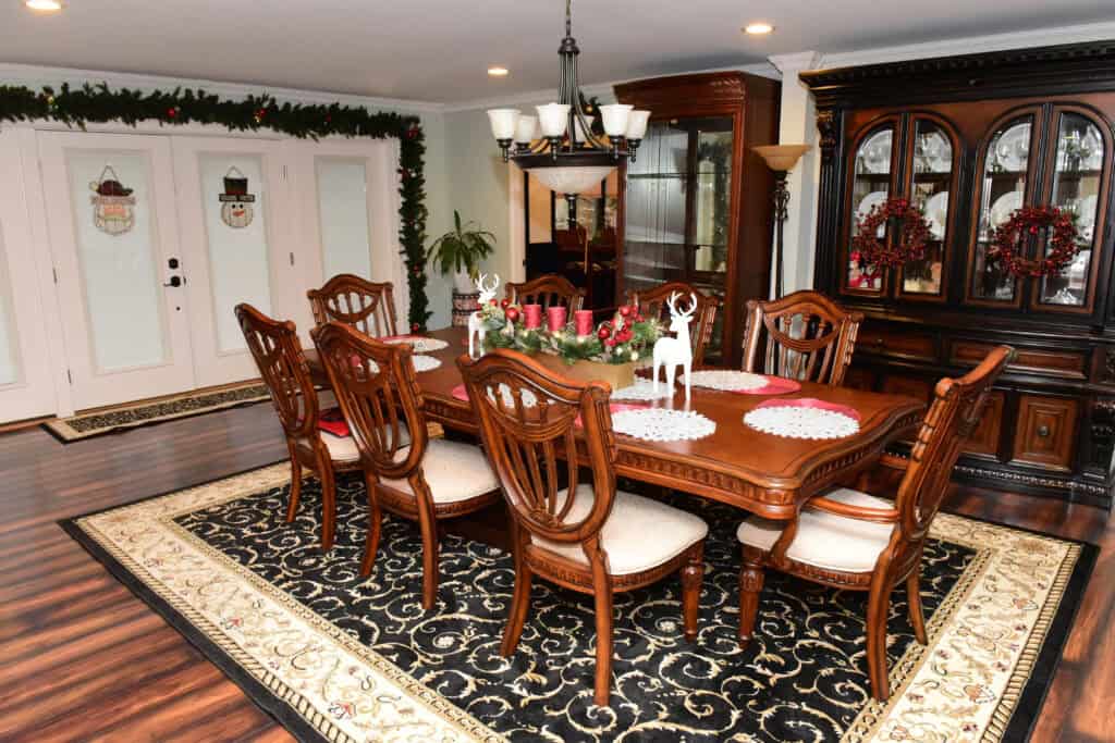 A formal dining room with a wooden table set for eight, holiday decorations including wreaths and candles, a decorative rug, china cabinet, and double doors adorned with snowman-themed decals and garland.