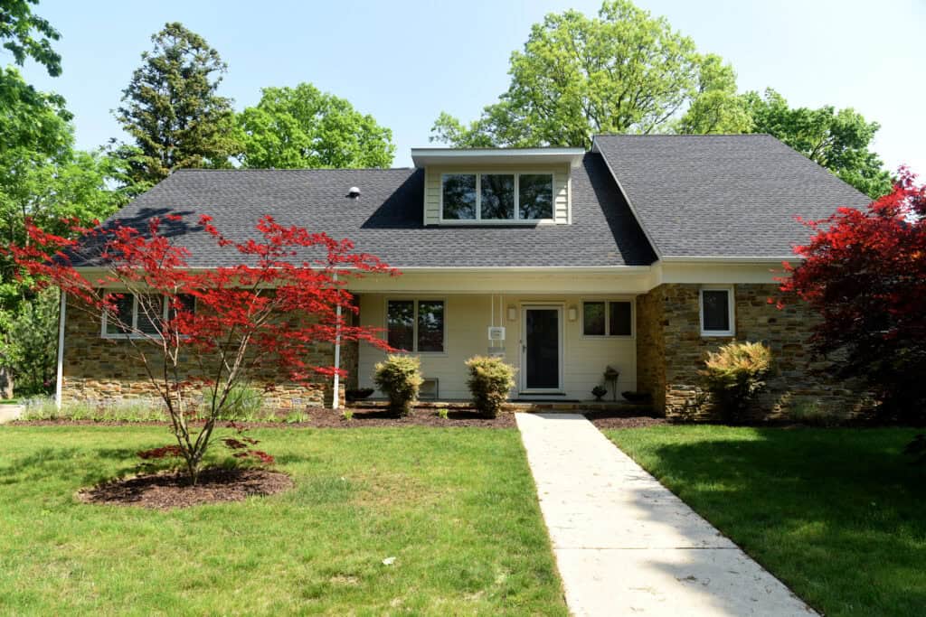 A tan house with a dark shingle roof and stone accents, surrounded by green grass, small red-leafed trees, and lush green trees in the background, under a clear, sunny sky. A sidewalk leads to the front door.