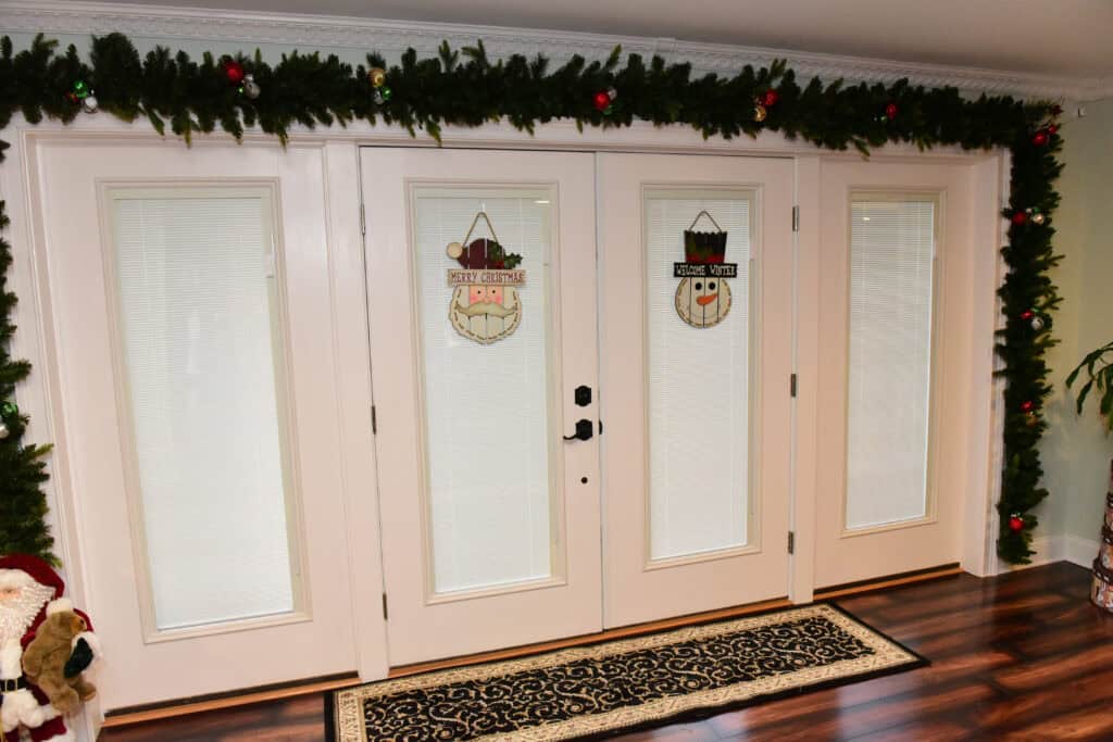 White double doors decorated with festive garland and ornaments along the top, with two holiday signs on the glass panels—one featuring a snowman and the other reading Merry Christmas. A doormat sits on the wooden floor.