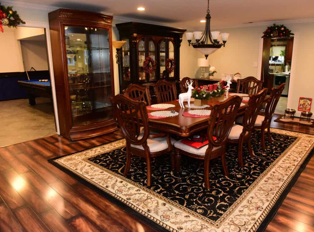A dining room with a large wooden table and eight chairs, set on a patterned rug. There are glass cabinets, Christmas wreaths, a chandelier, and a reindeer centerpiece. A pool table is visible in an adjacent room.