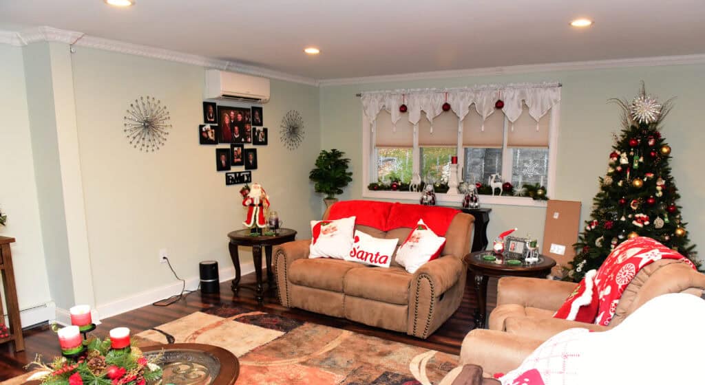 A cozy living room decorated for Christmas with a tree, red and white accents, a Santa pillow, holiday figurines on tables, and family photos on the wall. Wrapped gifts and candles add to the festive atmosphere.