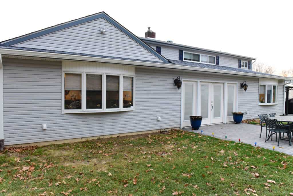 A backyard view of a house with light gray siding, white trim, a large set of windows, double glass doors, two wall lanterns, potted plants, a patio area with a table and chairs, and a grassy lawn with fallen leaves.