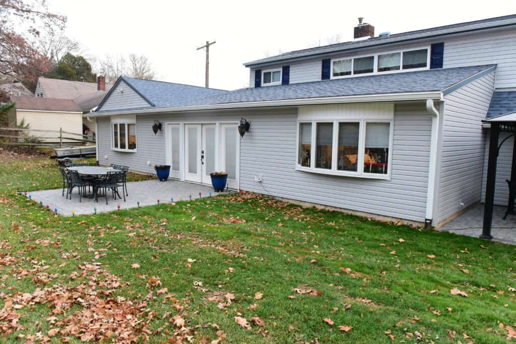 A light gray house with white trim features a patio with a black metal table and chairs, two blue planters by double doors, and a grassy yard with scattered fallen leaves.