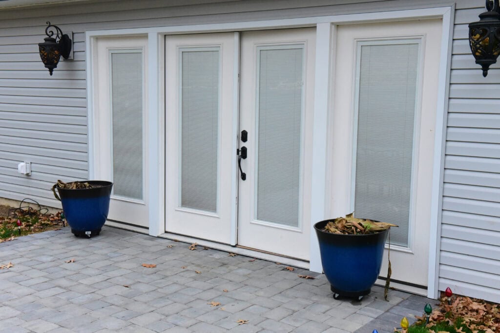 Double glass patio doors with white frames, flanked by two blue planters containing dried leaves, are set against light gray siding. Black lantern-style lights are mounted on both sides above the doors.
