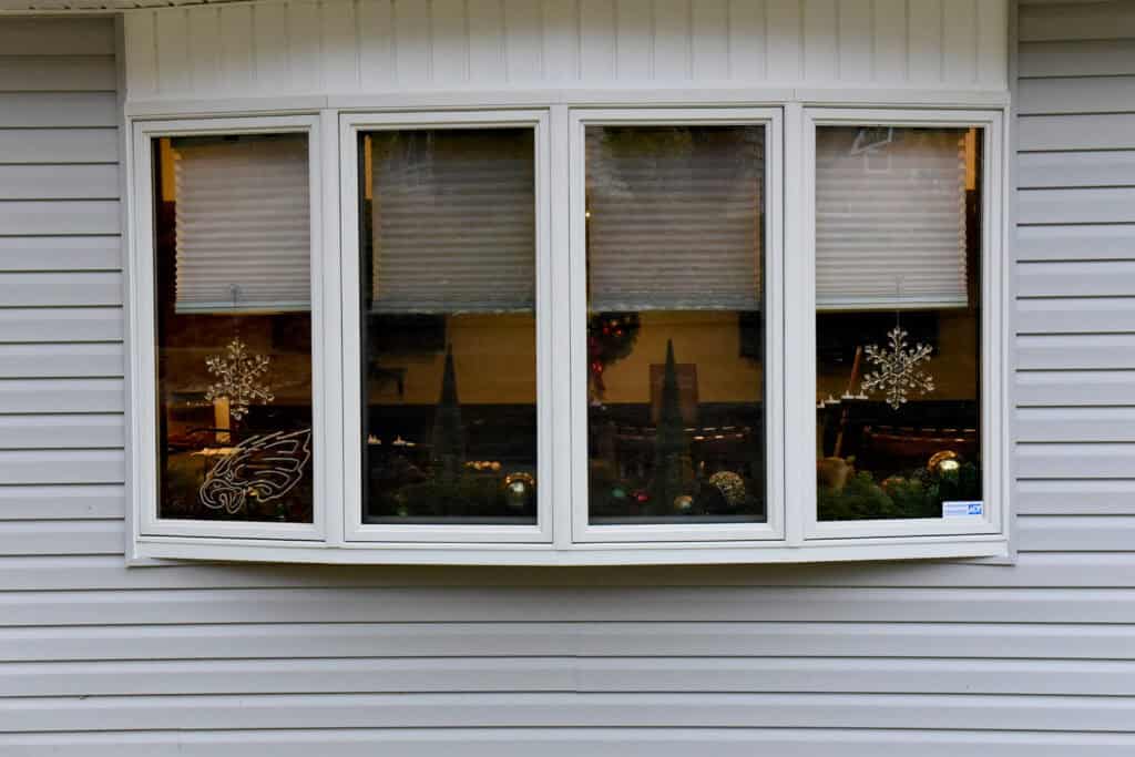 A white bay window with four glass panes and closed blinds, set in gray siding. Winter or holiday decorations, including snowflakes and ornaments, are visible inside.