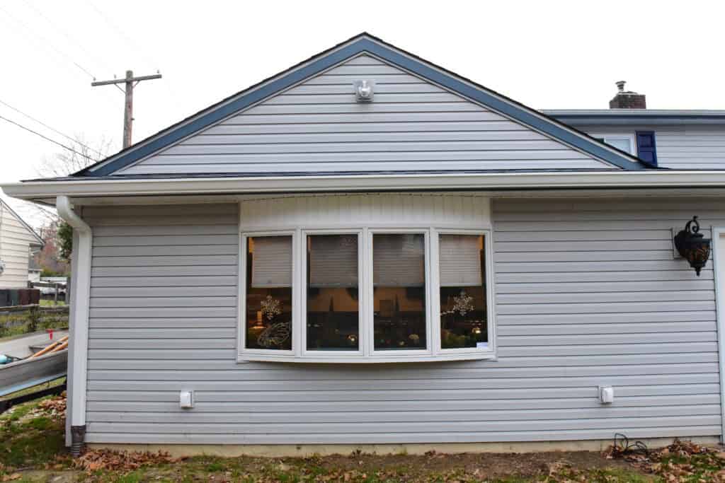 A gray house with white trim, featuring a large bay window. Warm lights and holiday decorations are visible inside. The ground in front has fallen leaves, and there is a black lantern on the right side of the house.
