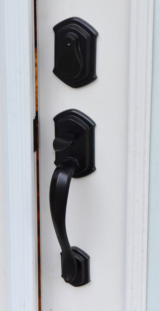 A close-up of a white door slightly ajar, showing a black handle and deadbolt lock on the edge.