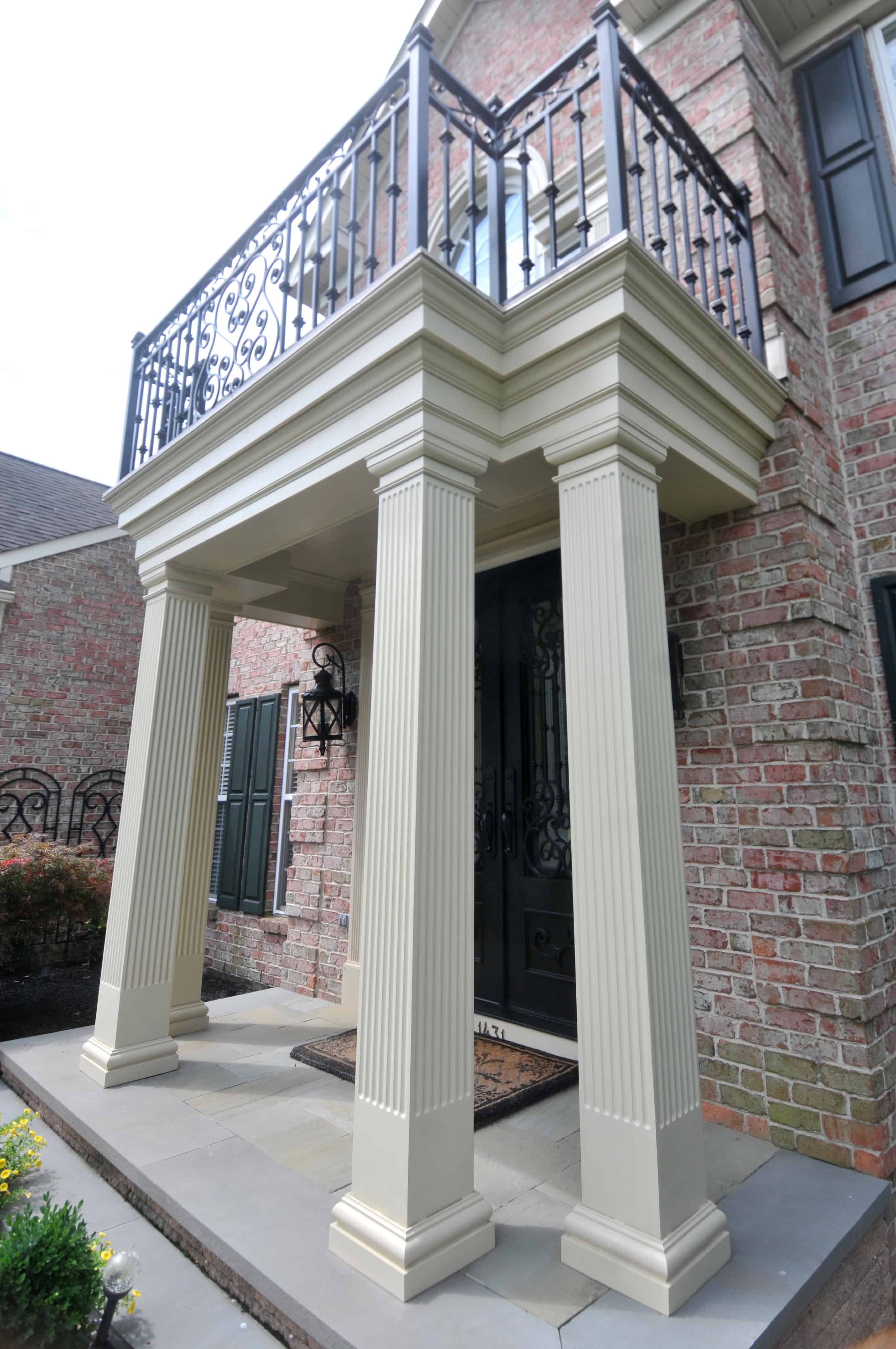 Angled view of a homes entrance featuring four fluted columns supporting a second-story balcony with ornate black metal railing, set against a brick exterior with black shutters.