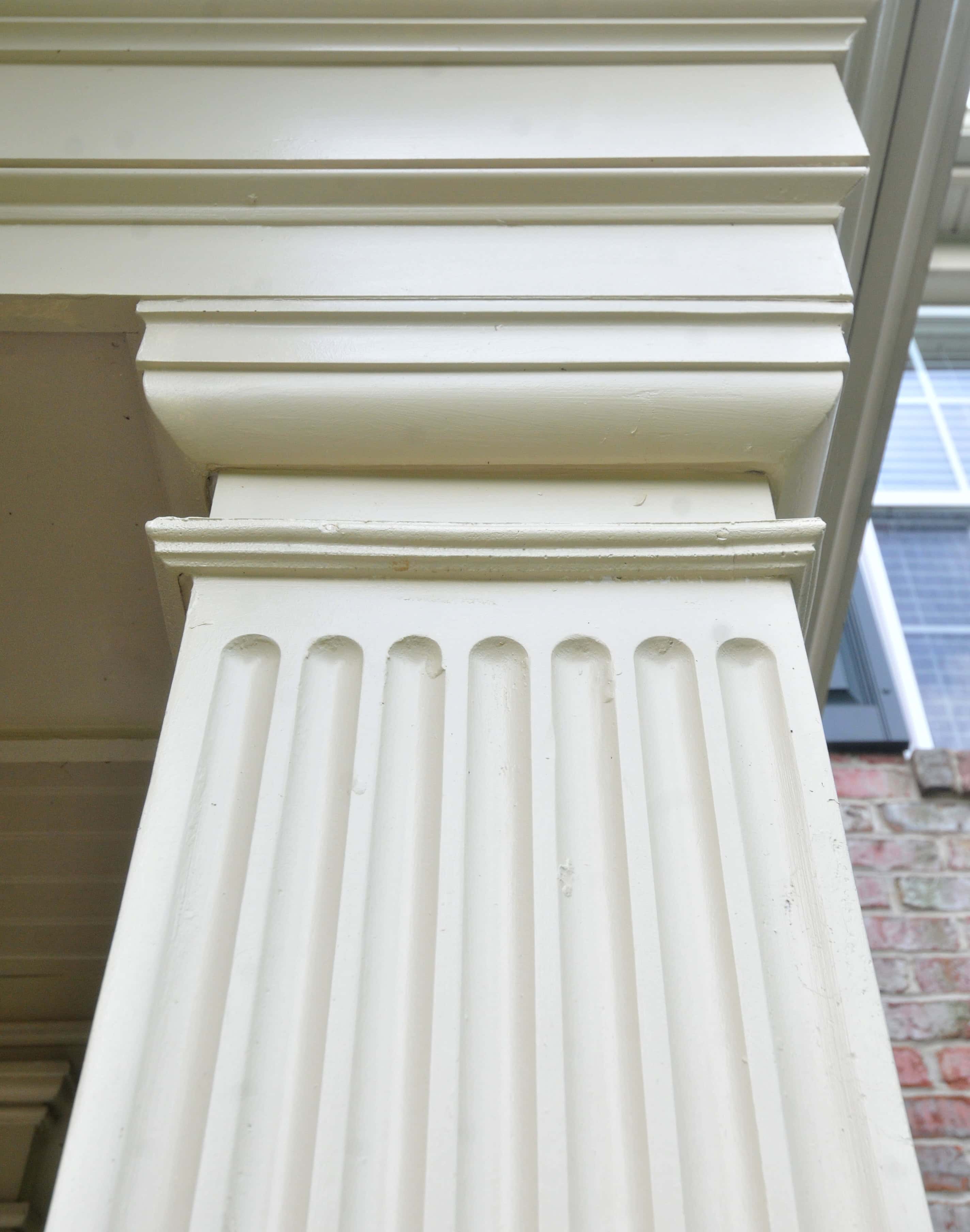 Close-up view of a white, fluted column with decorative molding at the top, supporting a structure. Windows and part of a brick wall are visible in the background.