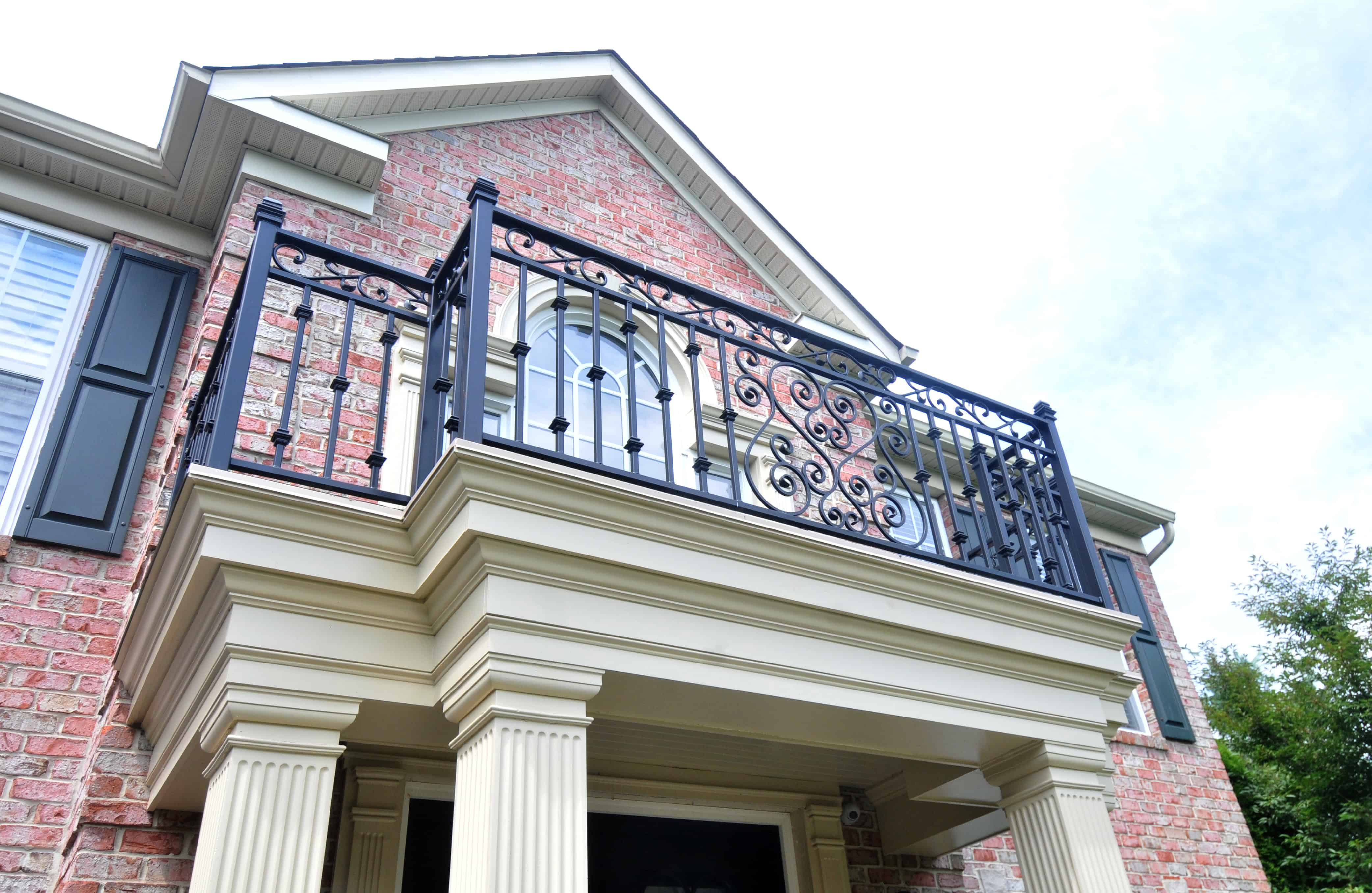 A close-up view of a house’s brick exterior with a decorative black wrought iron balcony railing above the entrance, featuring pillars, cream trim, and an arched window behind the railing.