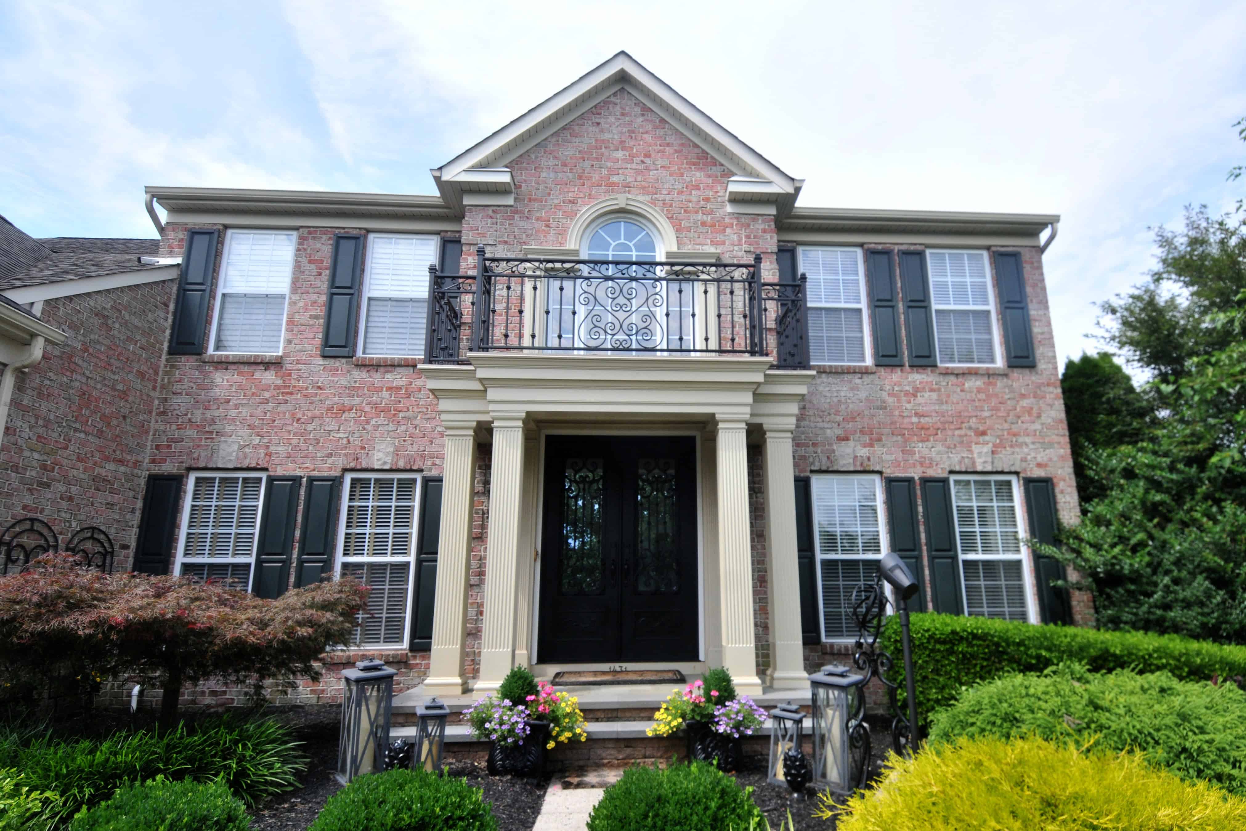A two-story brick house with black shutters, a small balcony over the main entrance, pillars framing the doorway, and a landscaped front yard with green bushes and colorful flowers.