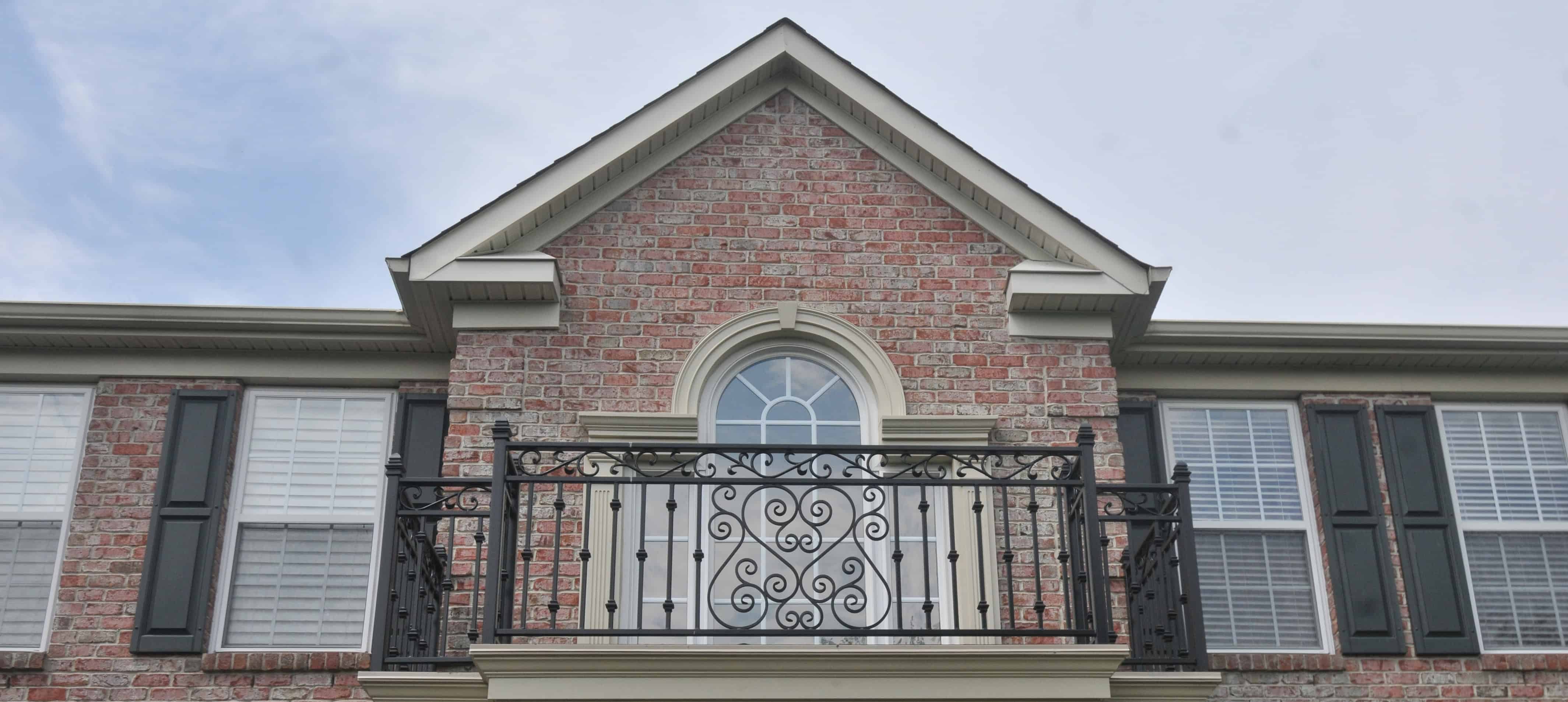A brick house with a decorative wrought iron balcony railing, arched window in the center, and white-trimmed windows with black shutters on either side, under a cloudy sky.