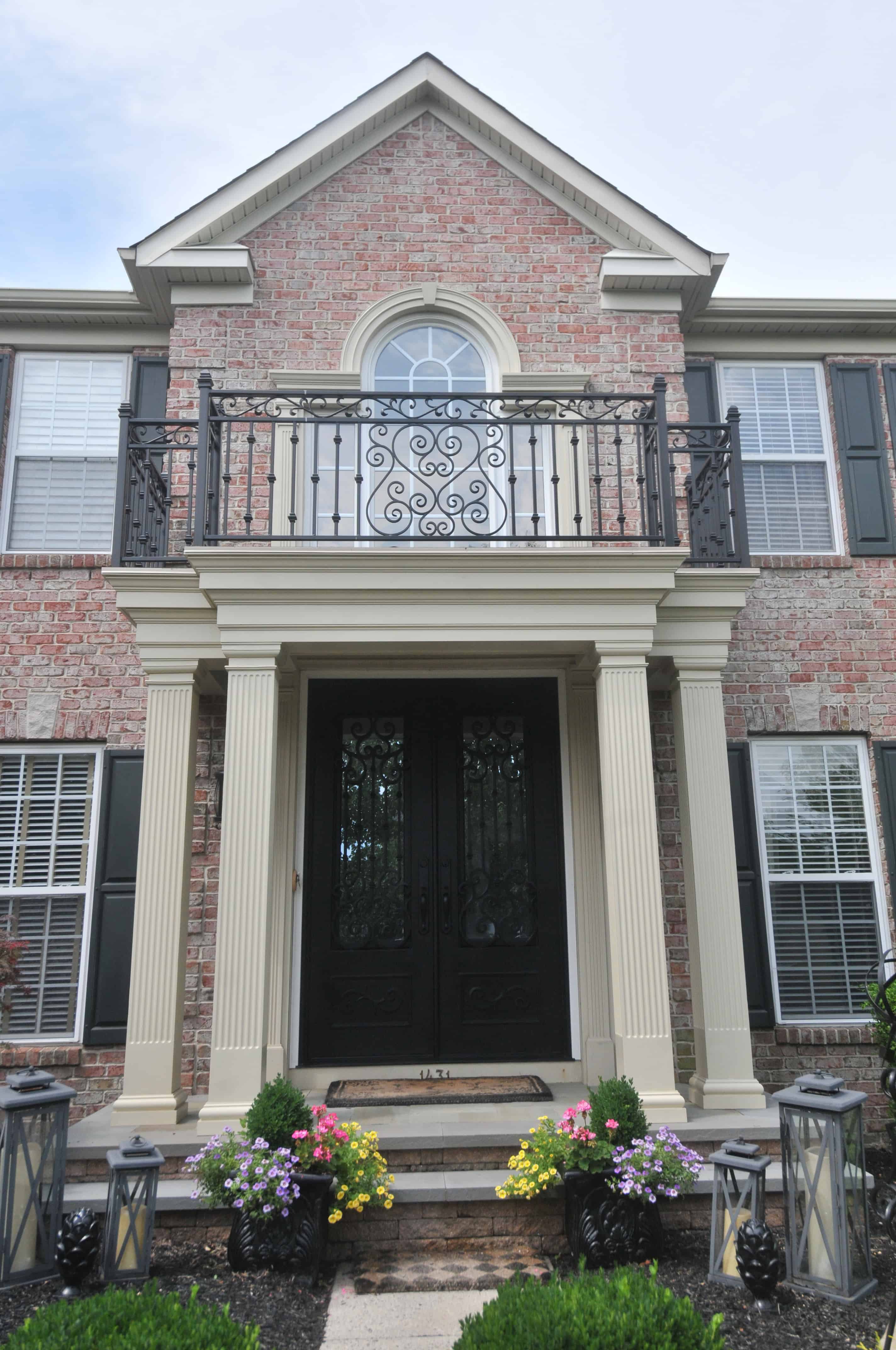 A two-story brick house with white columns, black double front doors, decorative iron balcony, and colorful flowers lining the entryway. Tall lanterns sit on either side of the walkway leading to the door.