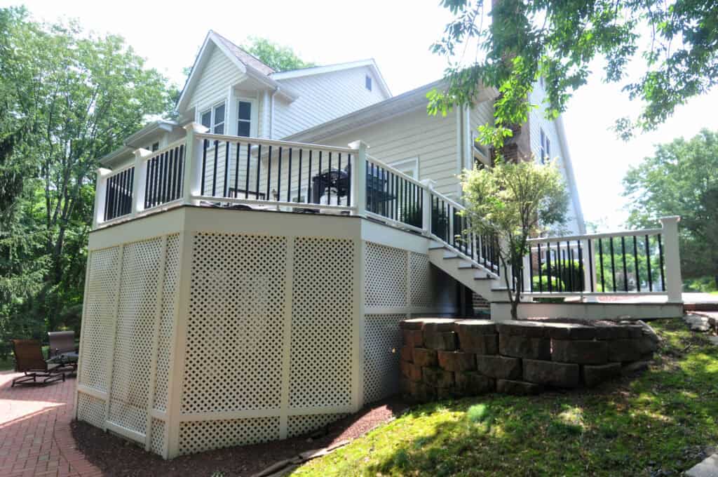 A large elevated deck with white lattice skirting, black railings, and stairs leading down to a patio surrounded by trees and a stacked stone retaining wall.