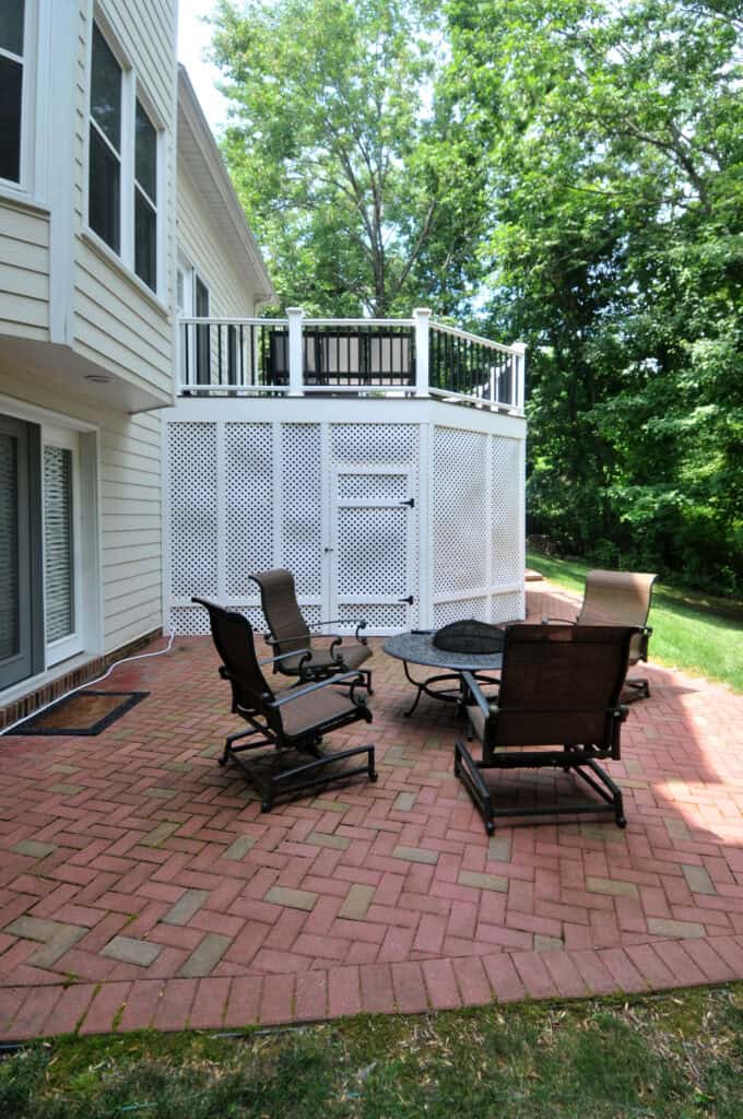A brick patio with four cushioned chairs around a metal fire pit, adjacent to a cream-colored house. Above, a white lattice enclosure supports a balcony with a railing. Green trees are visible in the background.