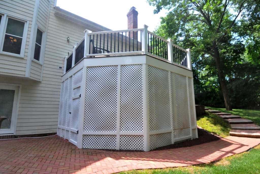 A raised deck attached to a house with white lattice panels underneath, black railing on top, and a brick patio and steps leading up a grassy yard beside it. Trees are visible in the background.