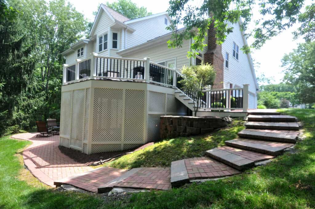 A two-story white house with a raised deck featuring black railings, stairs leading down to a brick patio, and a landscaped yard with stone steps and green grass. Trees surround the area, providing shade.