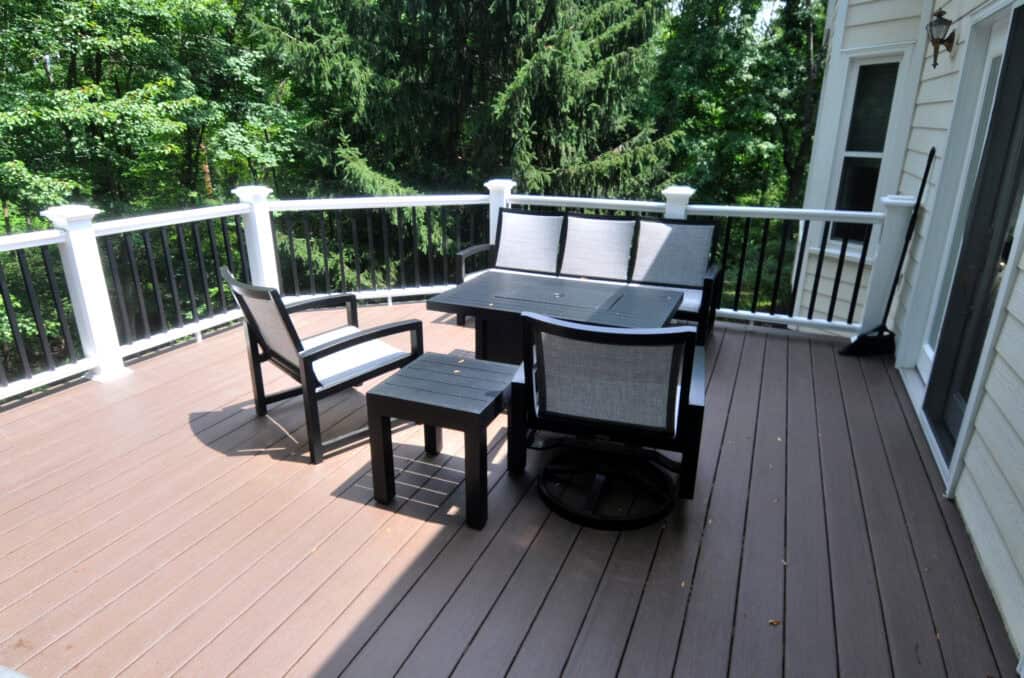 A wooden deck with black and white patio furniture, including a table, two chairs, and a bench, is surrounded by a white railing; green trees are visible in the background.
