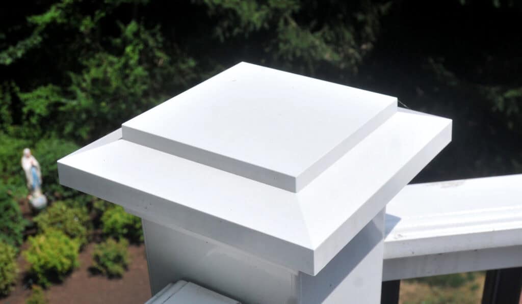 A close-up view of a white, square post cap on a fence or railing, with greenery and a small garden statue visible in the blurred background.