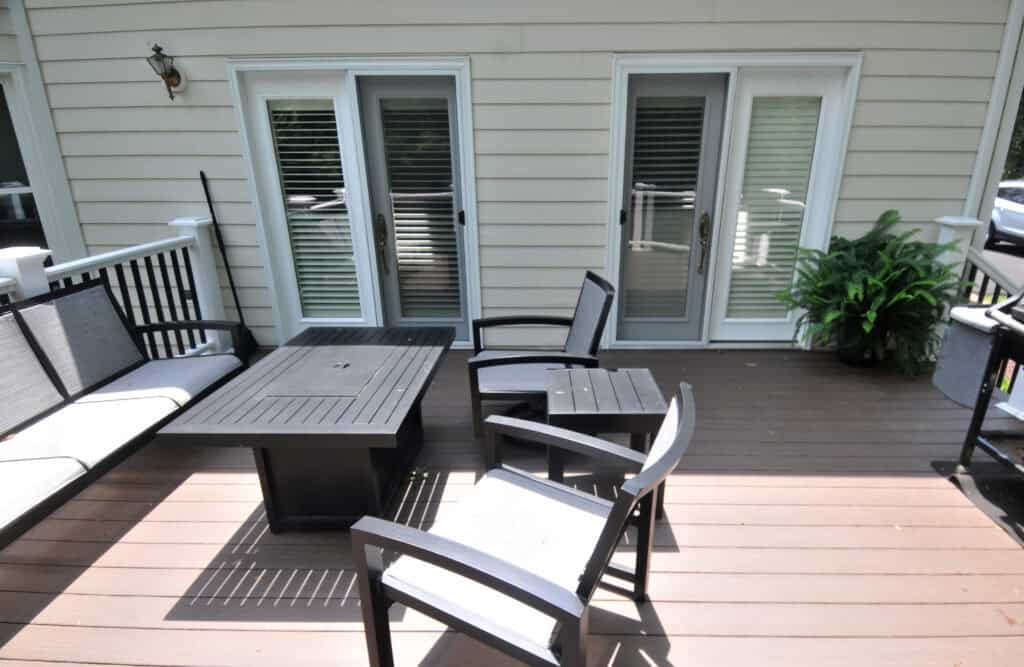 Outdoor patio with dark wooden seating, a matching table, and a white bench. The deck has brown flooring, a potted plant in the corner, and double glass doors with white shutters leading inside.