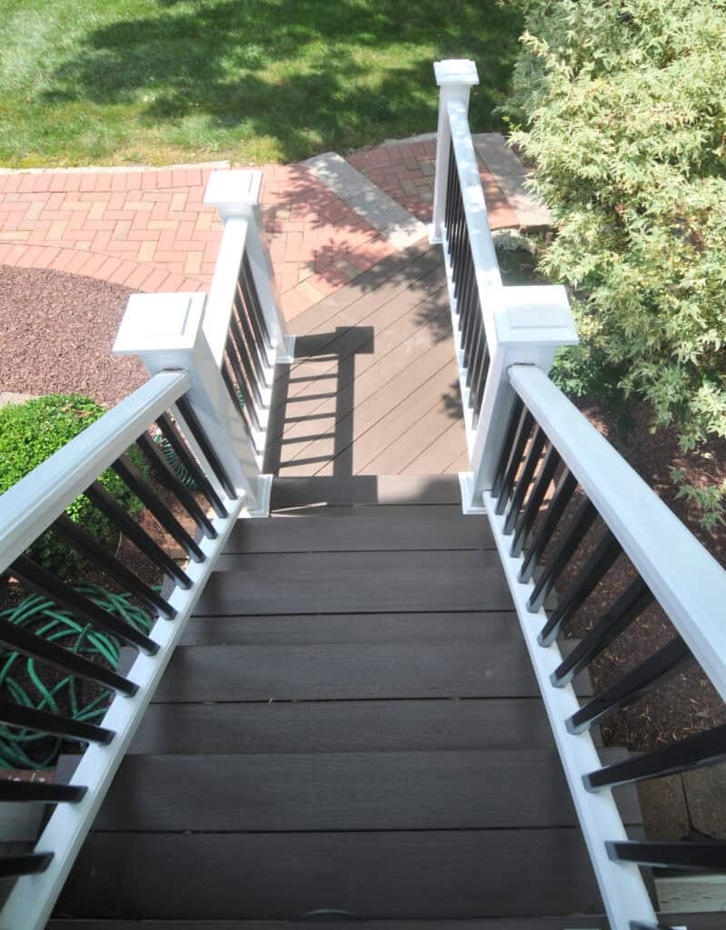 View looking down a small outdoor staircase with dark brown steps and white railings, leading to a garden area with brick and stone paths and green foliage.