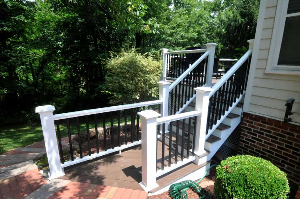 A wooden deck with white and black railing leads to stairs, surrounded by green bushes and trees. The deck is attached to a brick house with a garden hose on the ground nearby.