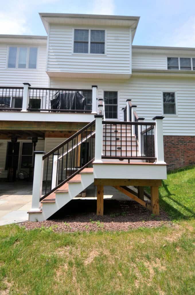 A white house with a two-level deck and black railings. Wooden steps lead from the upper deck to the grassy backyard. Part of the patio and basement area are visible beneath the deck.
