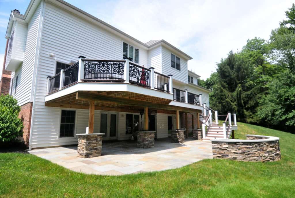 A two-story white house with a large stone patio, covered area supported by stone columns, upper balcony with decorative railing, and a circular stone fire pit in a grassy backyard.