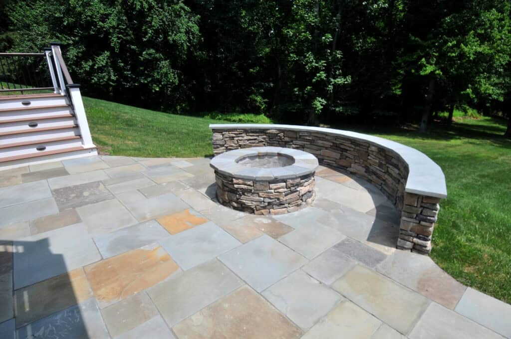 A stone patio with a built-in circular stone fire pit and a curved stone bench, surrounded by grass and trees in a backyard.