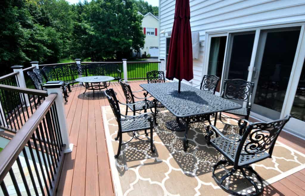A wooden backyard deck with ornate black metal patio furniture, including a dining table with chairs, a smaller round table, and an umbrella. The deck overlooks a grassy yard with trees and a white house nearby.