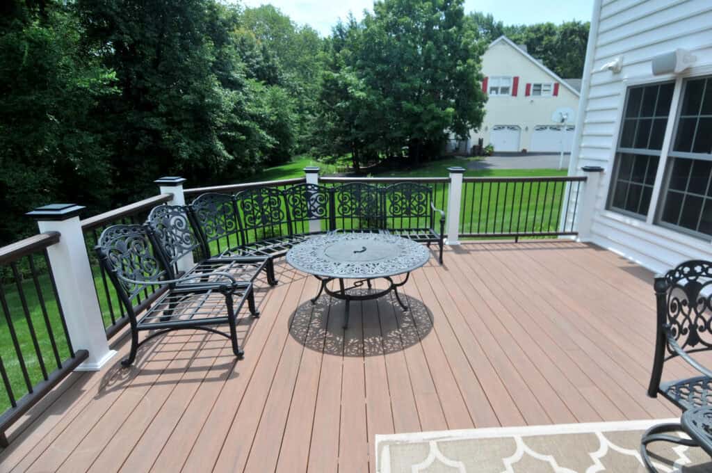 A spacious wooden deck with ornate black metal patio furniture, including a round table and benches, overlooks a green lawn and trees, with a white house and a three-car garage in the background.