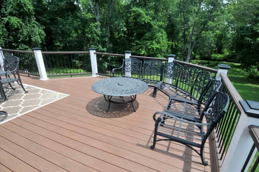 A backyard deck with brown flooring, black metal railings, ornate black metal benches, a round metal table in the center, and a beige patterned outdoor rug, surrounded by lush green trees.