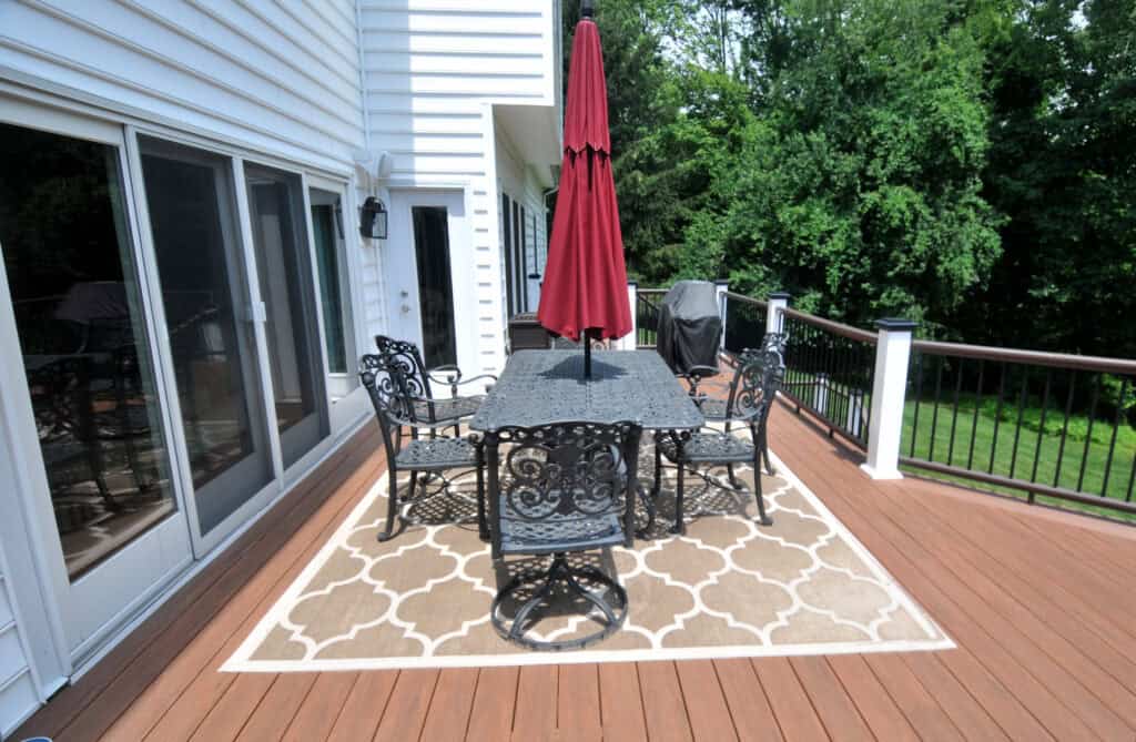 A patio with a black metal table and chairs, a red umbrella, and a patterned outdoor rug sits on a wooden deck next to a white house, overlooking a green, tree-filled yard.