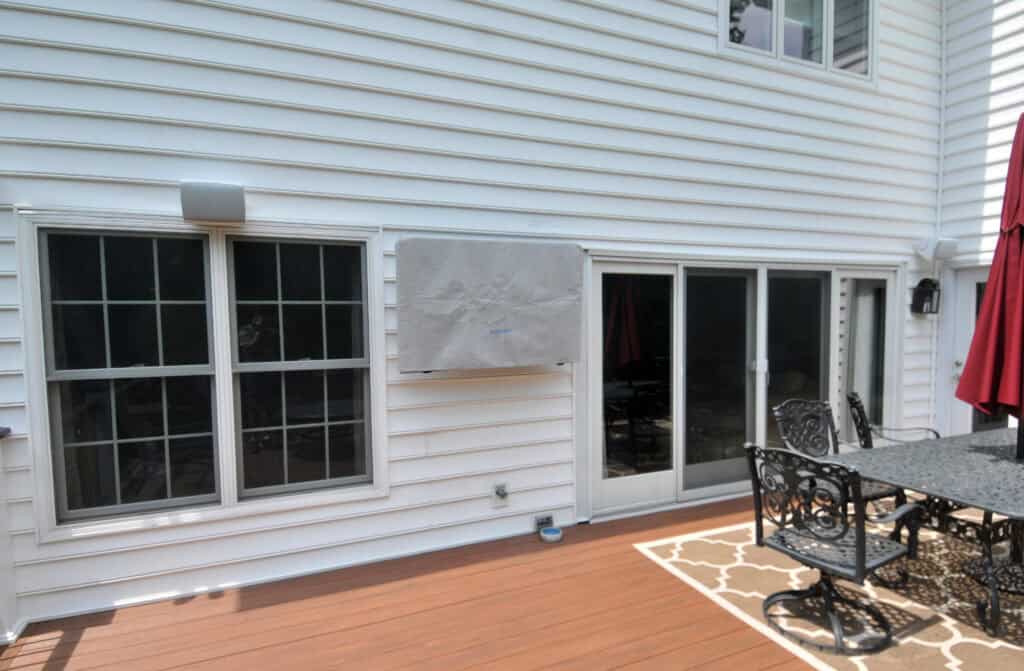 A white house with two large windows and a glass sliding door opening onto a wooden deck with black metal patio furniture, a rug, and a red umbrella. An outdoor TV is covered and mounted on the wall.