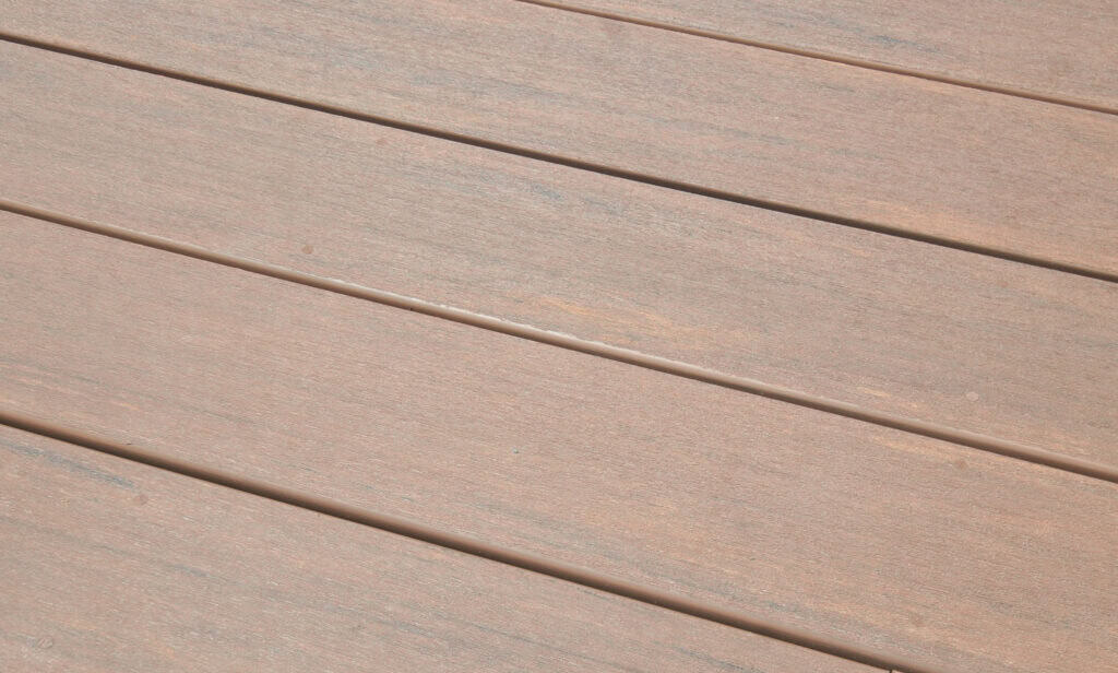 Close-up view of brown wooden planks laid side by side, forming a smooth and even surface, possibly part of a deck or floor. The wood grain and subtle color variations are visible.