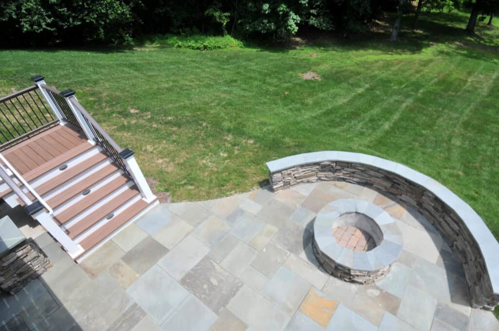 Patio with stone tiles features a circular stone fire pit and a short staircase with white railings leading down to a grassy backyard surrounded by trees.