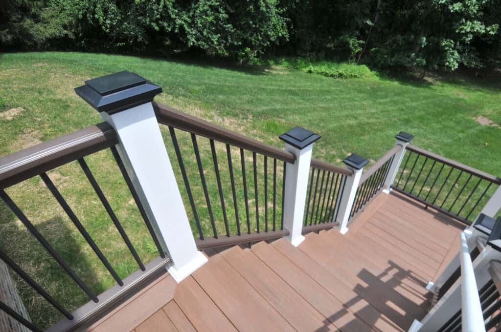 A view looking down wooden outdoor stairs with brown railings and white posts, leading to a green grassy lawn bordered by trees.
