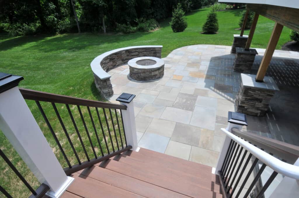 View from stairs leading to a backyard patio with stone pavers, a built-in curved stone bench, and a circular fire pit, surrounded by a green lawn and trees in the background.