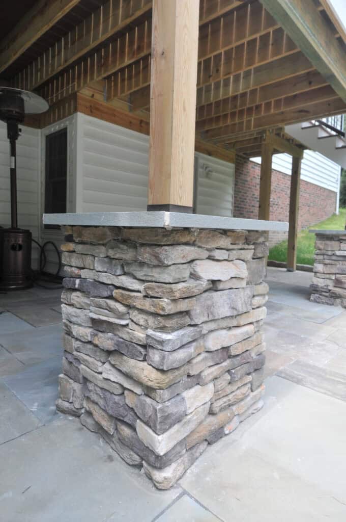 A wooden post supported by a rectangular stone pillar on a patio, with a patio heater nearby and a partially visible brick wall and white siding in the background.