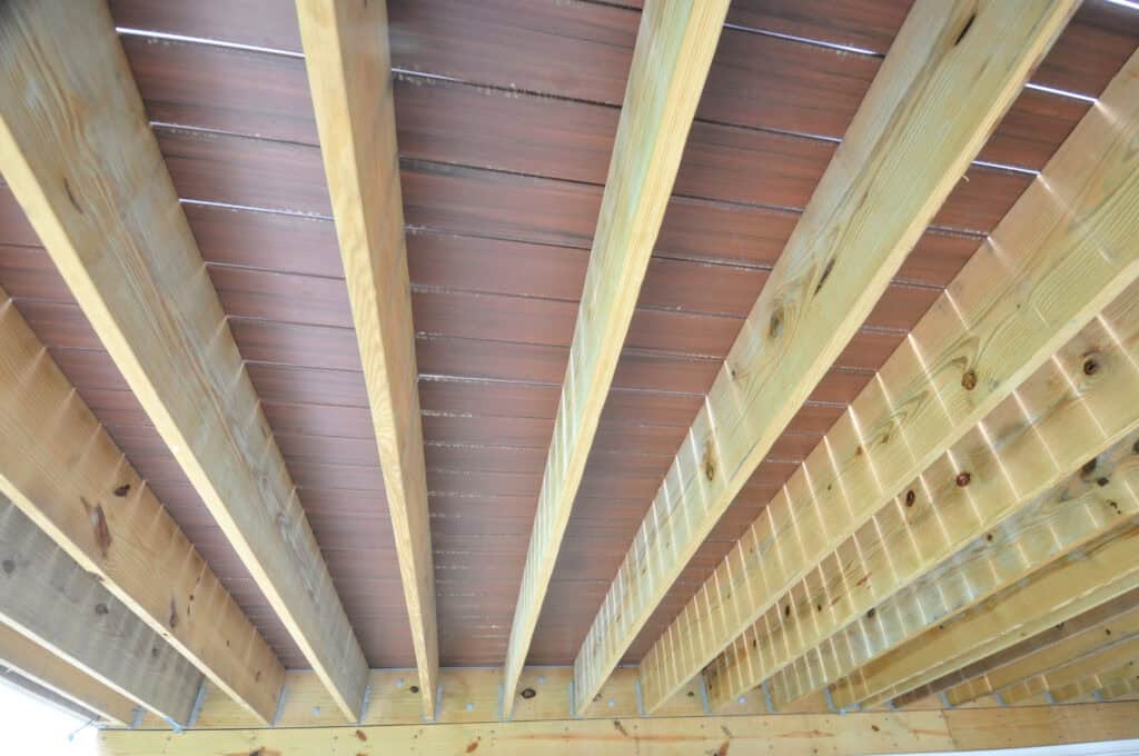 Close-up view of the underside of a wooden deck, showing parallel wooden beams and reddish-brown decking boards above, arranged in a repeating pattern.