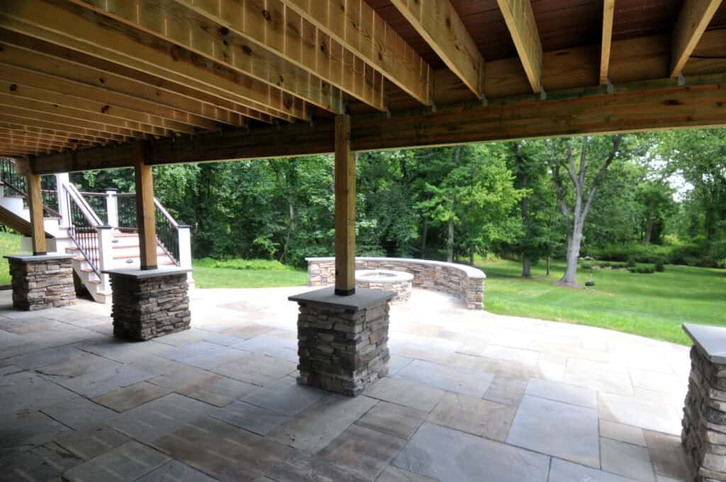 View from under a wooden deck with exposed beams, stone pillars, and a paved patio, overlooking a green, tree-filled backyard. Steps lead up to the deck on the left side.