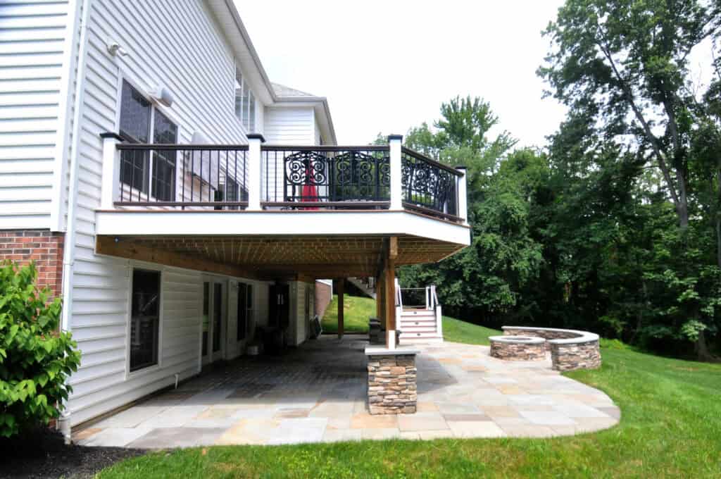 A two-story house with a white exterior and brick accents features an elevated, railed deck supported by stone columns. Below, there is a covered patio with stone flooring, next to a landscaped yard and curved stone seating wall.